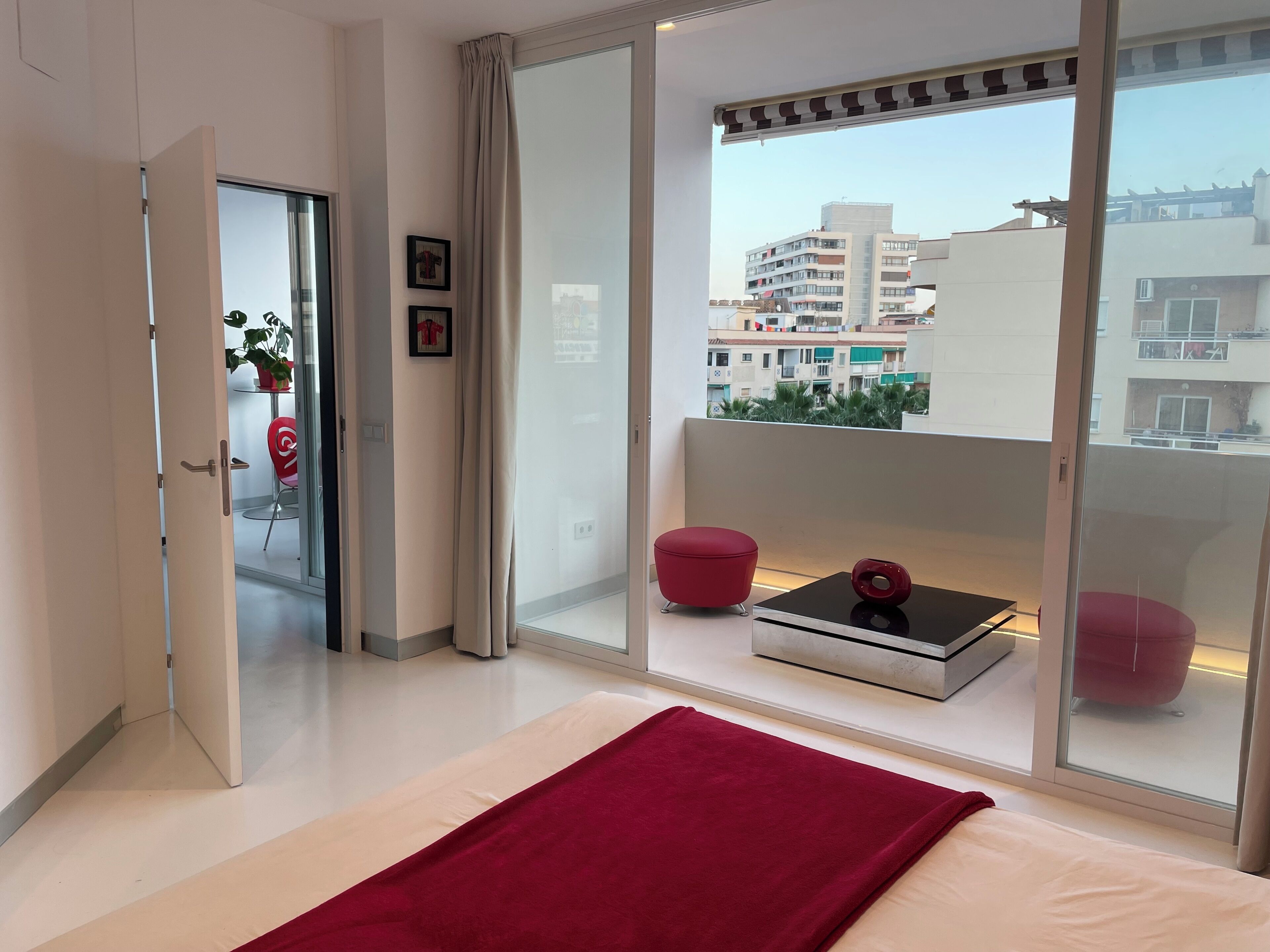 A modern bedroom with a large sliding glass door leading to a balcony, a bed with a red throw, and a small table with a decorative item.