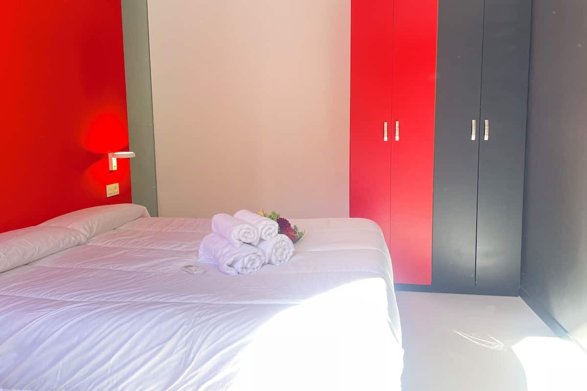 A hotel room with a bed, a red wardrobe, and a nightstand with a lamp.
