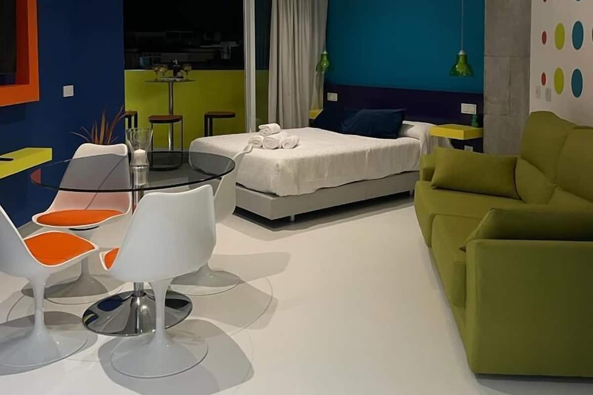 A modern hotel room with a bed, a green sofa, a dining table with chairs, and a ceiling light.