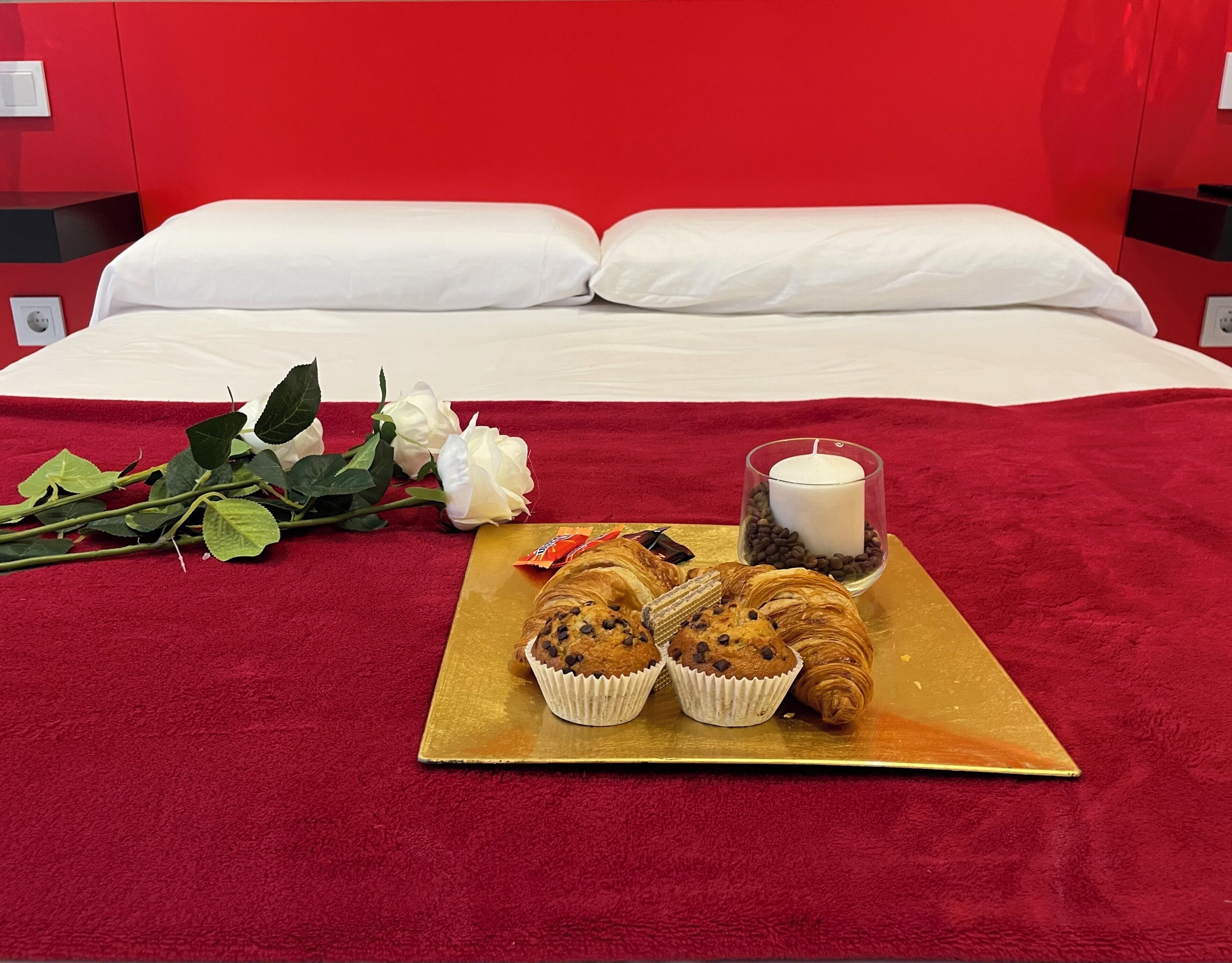 A bed with white linens, a red blanket, a tray with croissants and muffins, a candle, and a bouquet of white roses.
