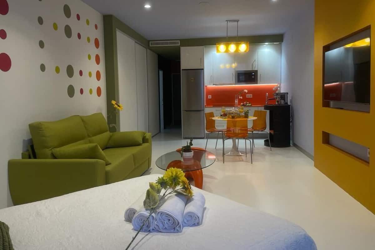 A modern hotel room with a green sofa, a dining table with orange chairs, a kitchen area with a microwave, and a TV mounted on the wall.