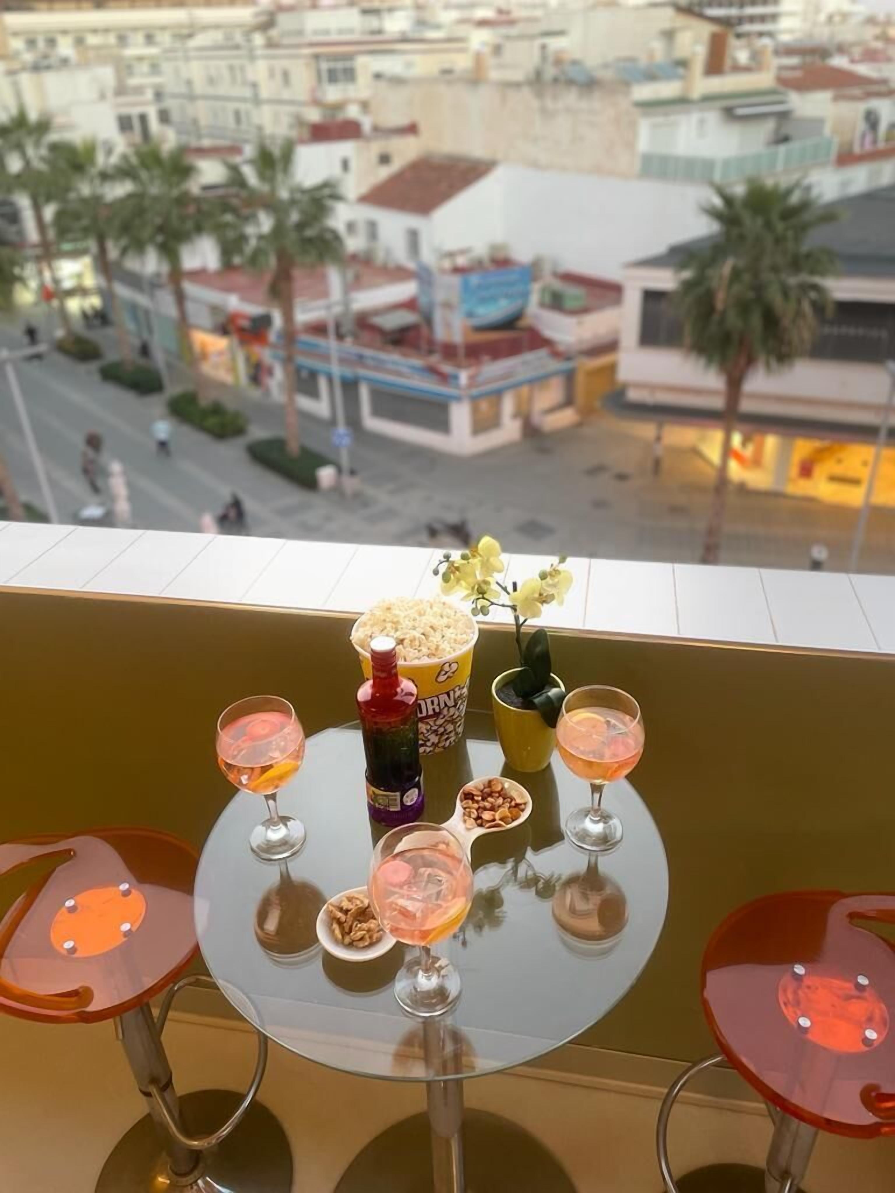 Un balcon avec une table dressée pour prendre des boissons et grignoter, donnant sur un paysage urbain avec des immeubles et des palmiers.