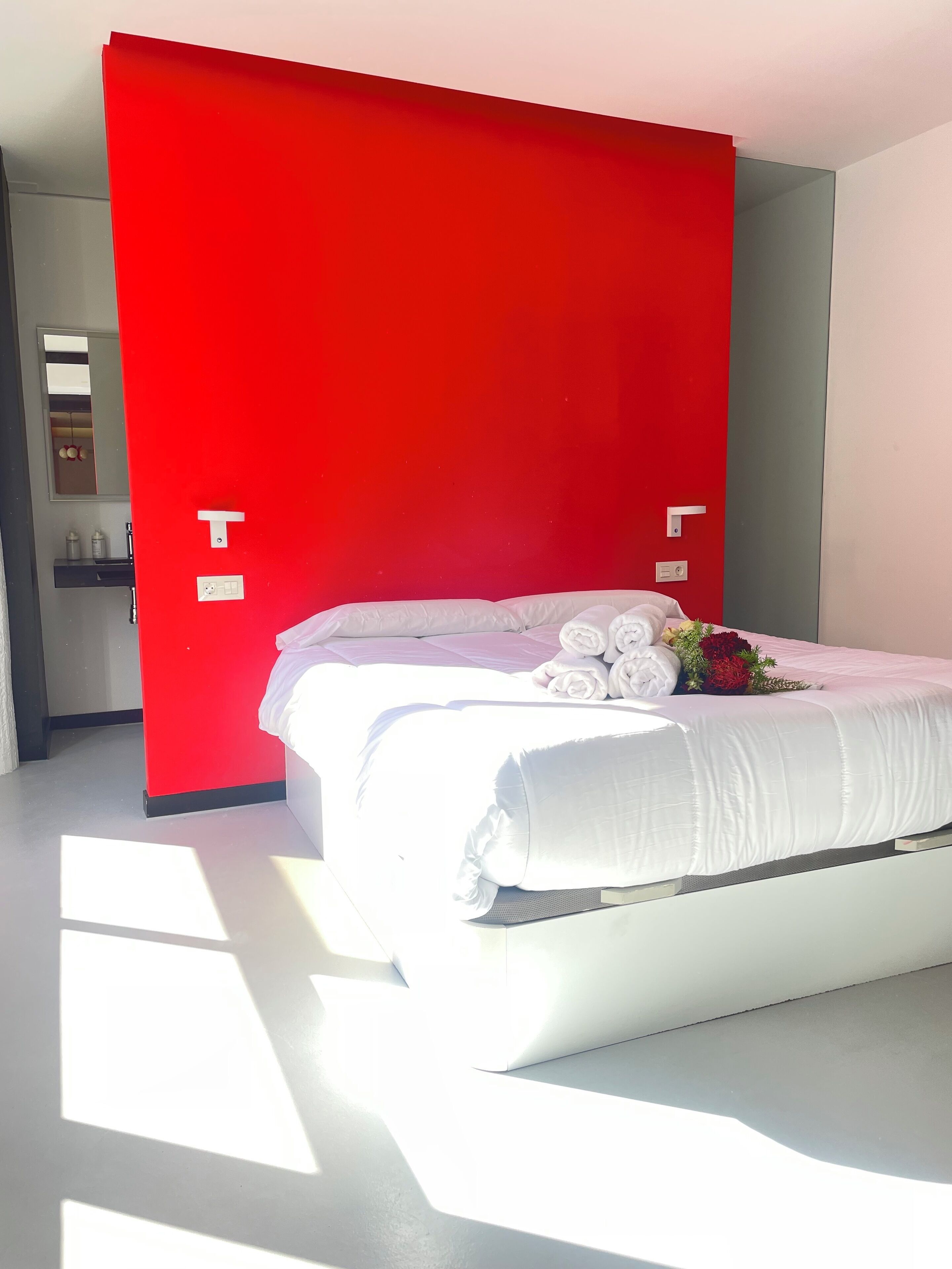 A modern hotel room with a large bed, white bedding, and a red accent wall.