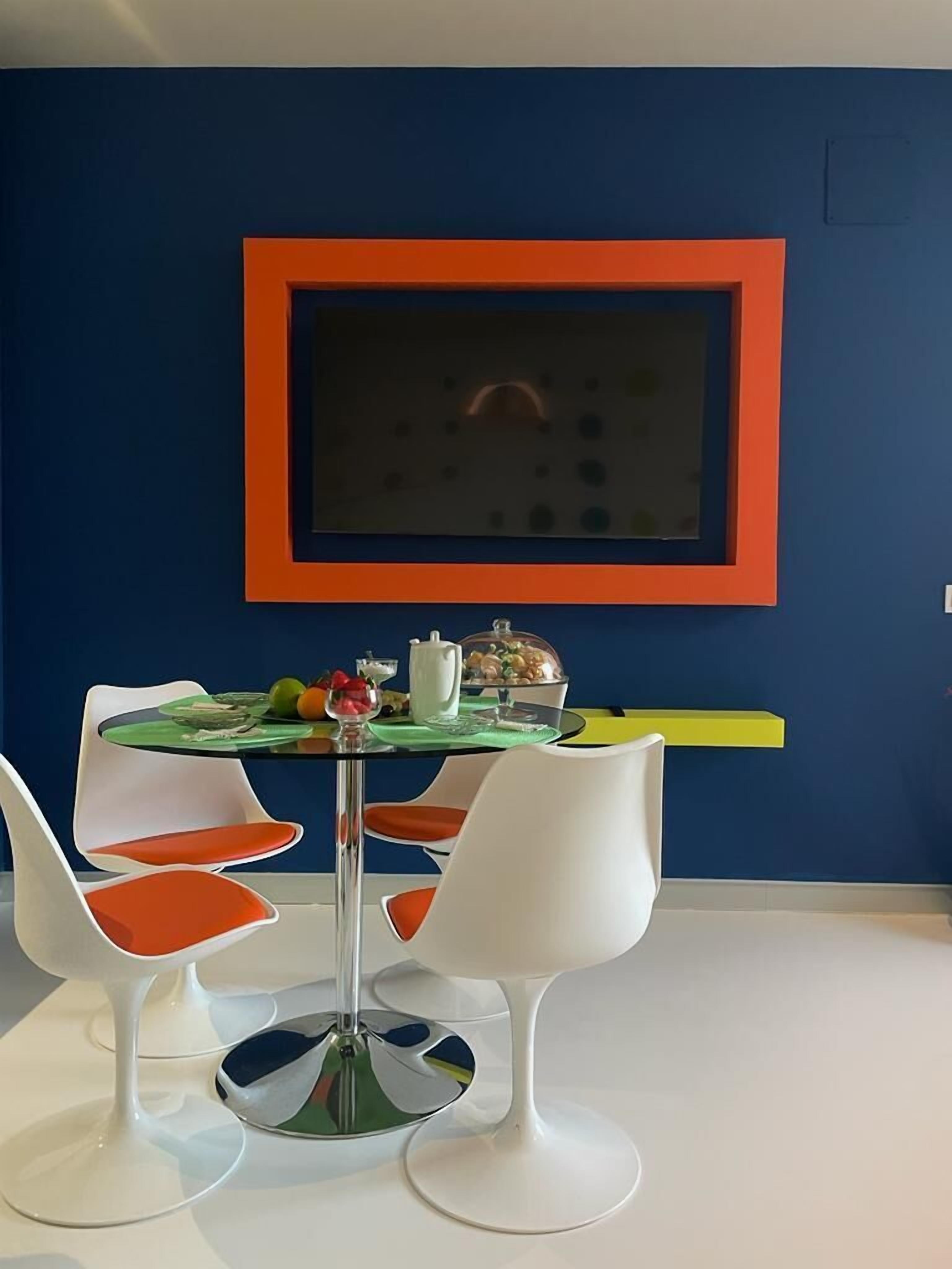A dining area with a round table, white chairs with orange cushions, and a blue wall with a red-framed picture.