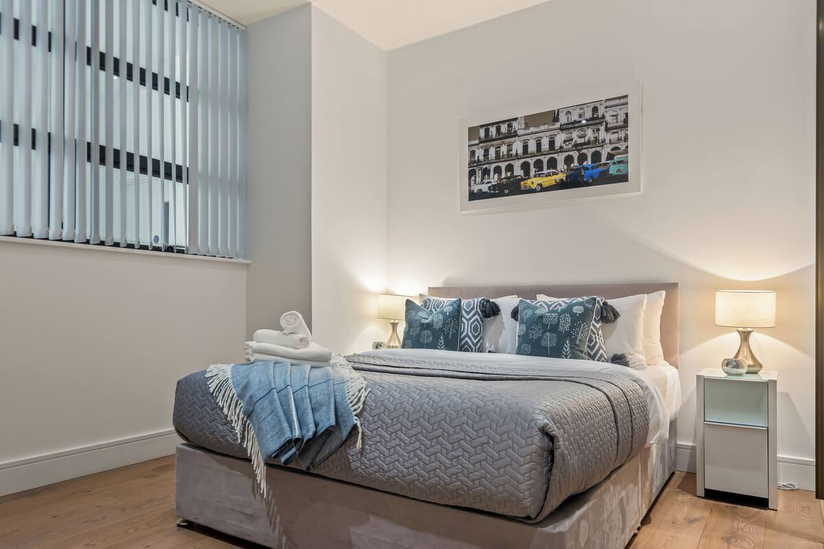 A neatly arranged bedroom with a bed, bedside lamps, a window with blinds, and a wall-mounted artwork.