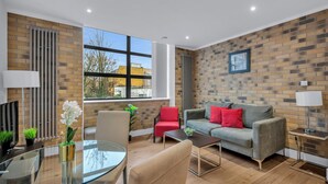 A modern living room with a glass coffee table, a grey sofa with red cushions, a red chair, and a brick-patterned wall. - Regent's Park Serviced Apartments (London)