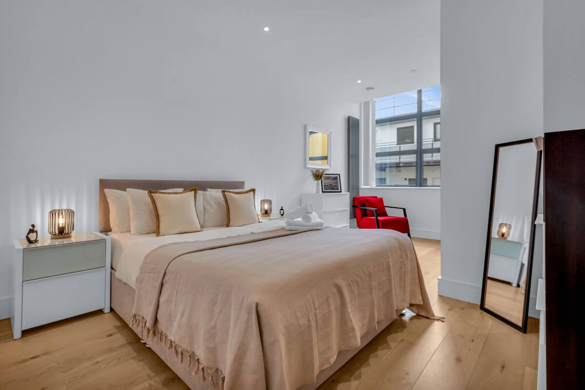 A modern hotel room with a large bed, bedside tables, a red chair, and a large window.