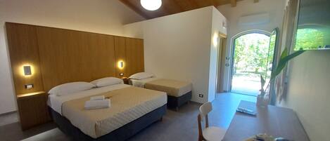 A hotel room with a large bed, bedside tables, a desk with a chair, a kettle, and a view of greenery outside.