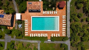 Μια αεροφωτογραφία ενός θέρετρου με μια κεντρική πισίνα, η οποία περιβάλλεται από ξαπλώστρες, μια σκεπαστή περιοχή με πλακάκια στην οροφή και έναν κήπο.