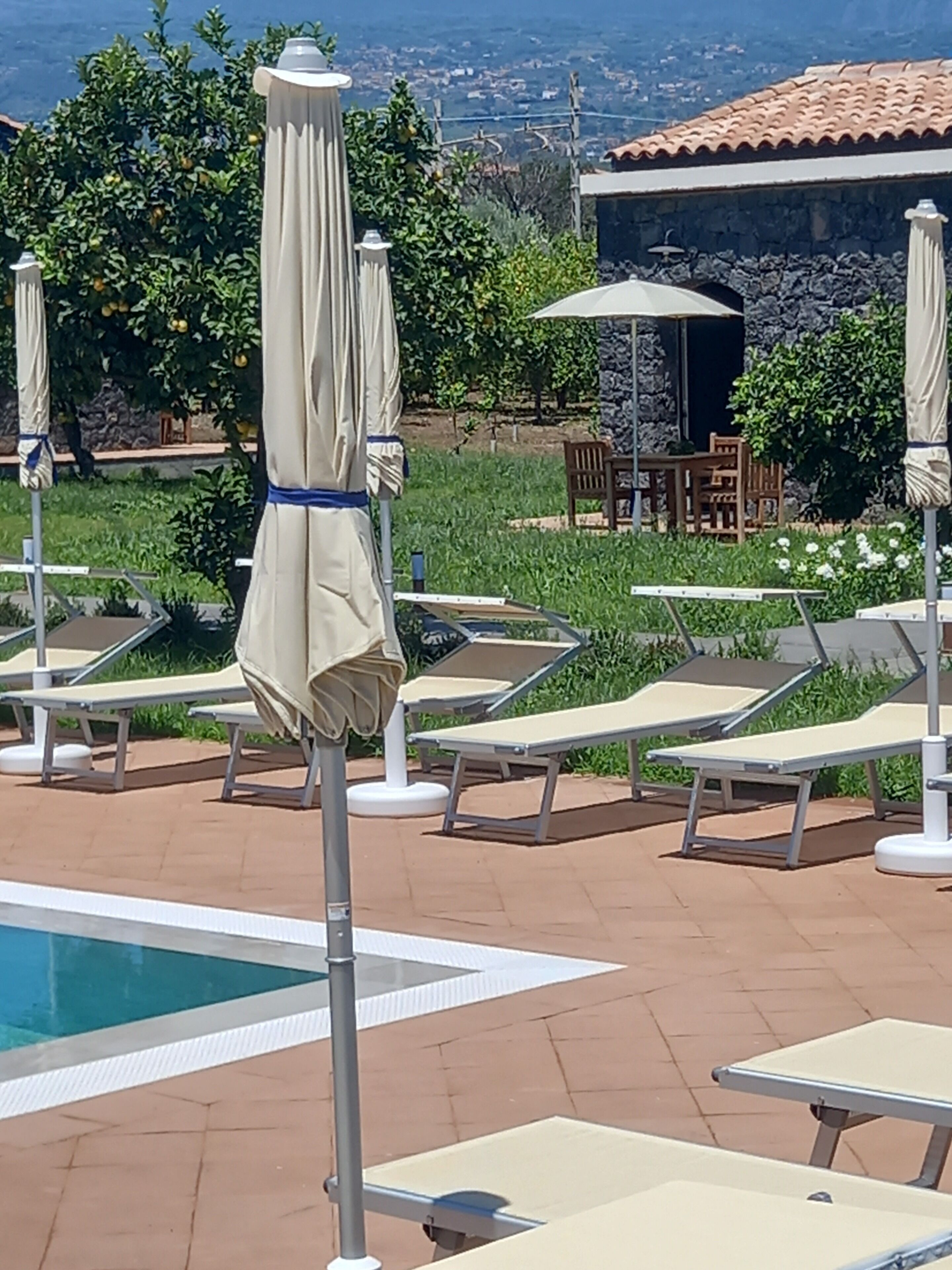 Zona junto a la piscina con sillones y sombrillas, un edificio de piedra al fondo y vistas del paisaje.