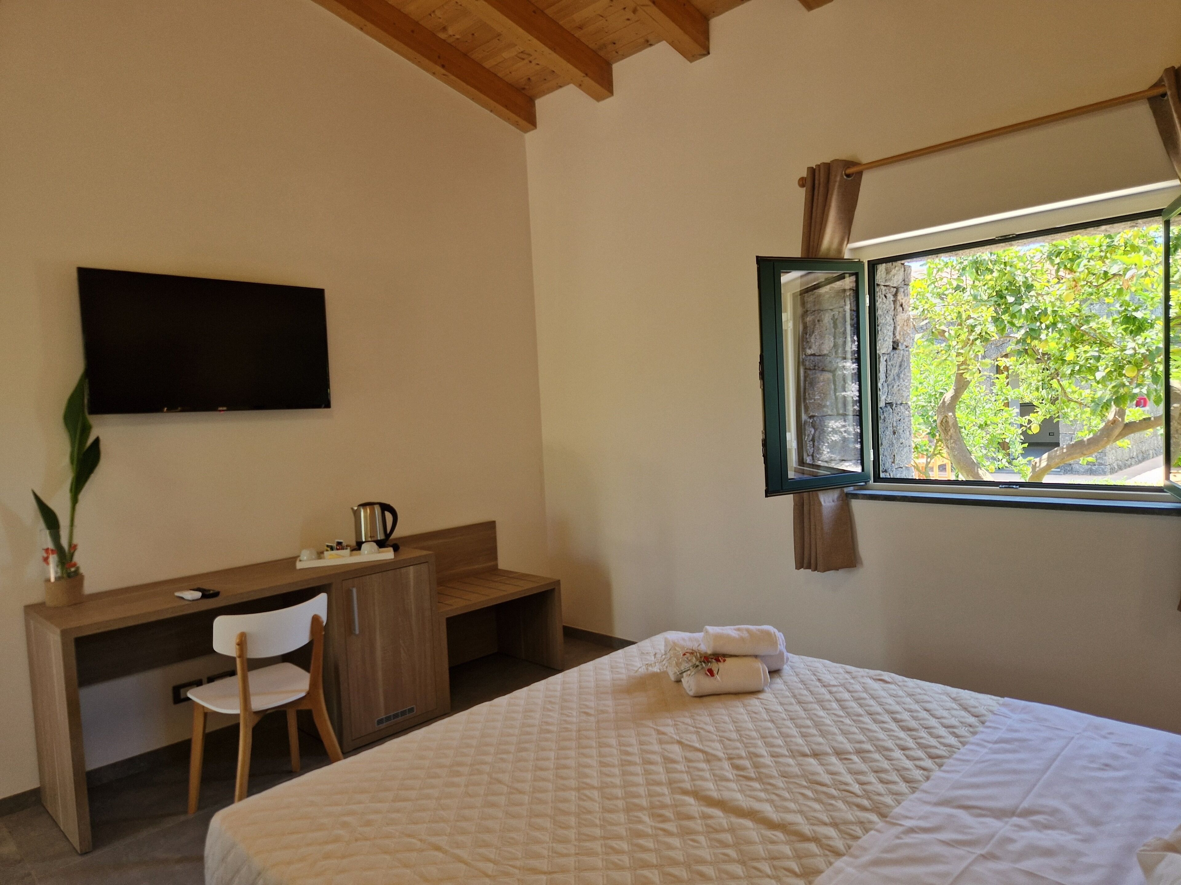 A bedroom with a bed, a desk with a chair, a television, and a window with a view of greenery.