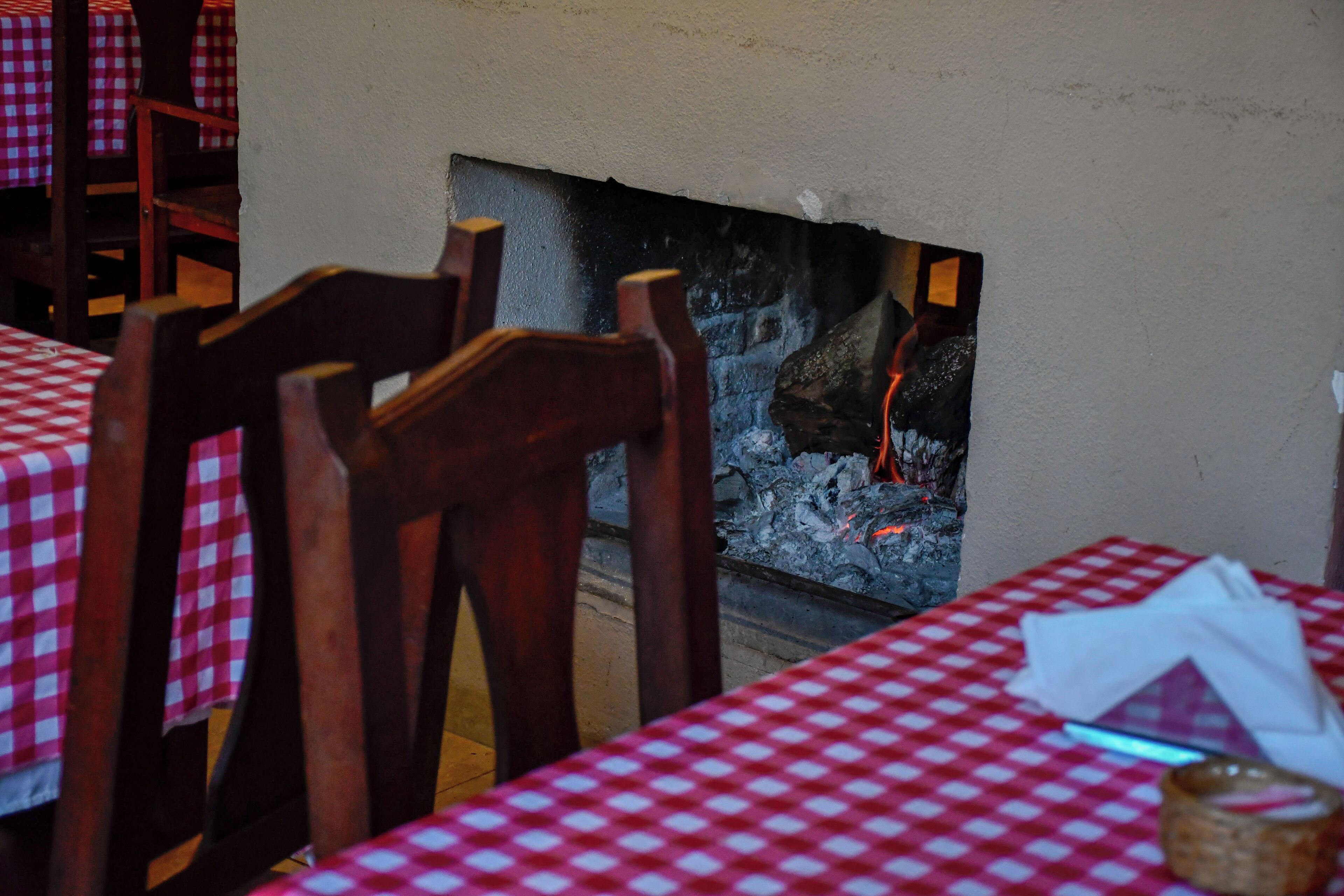Interior de restaurante con manteles a cuadros, sillas de madera y una chimenea con leña a la vista.