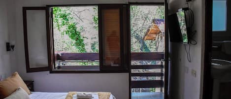 A hotel room with a bed, a TV, a window with a view of trees, and a balcony.