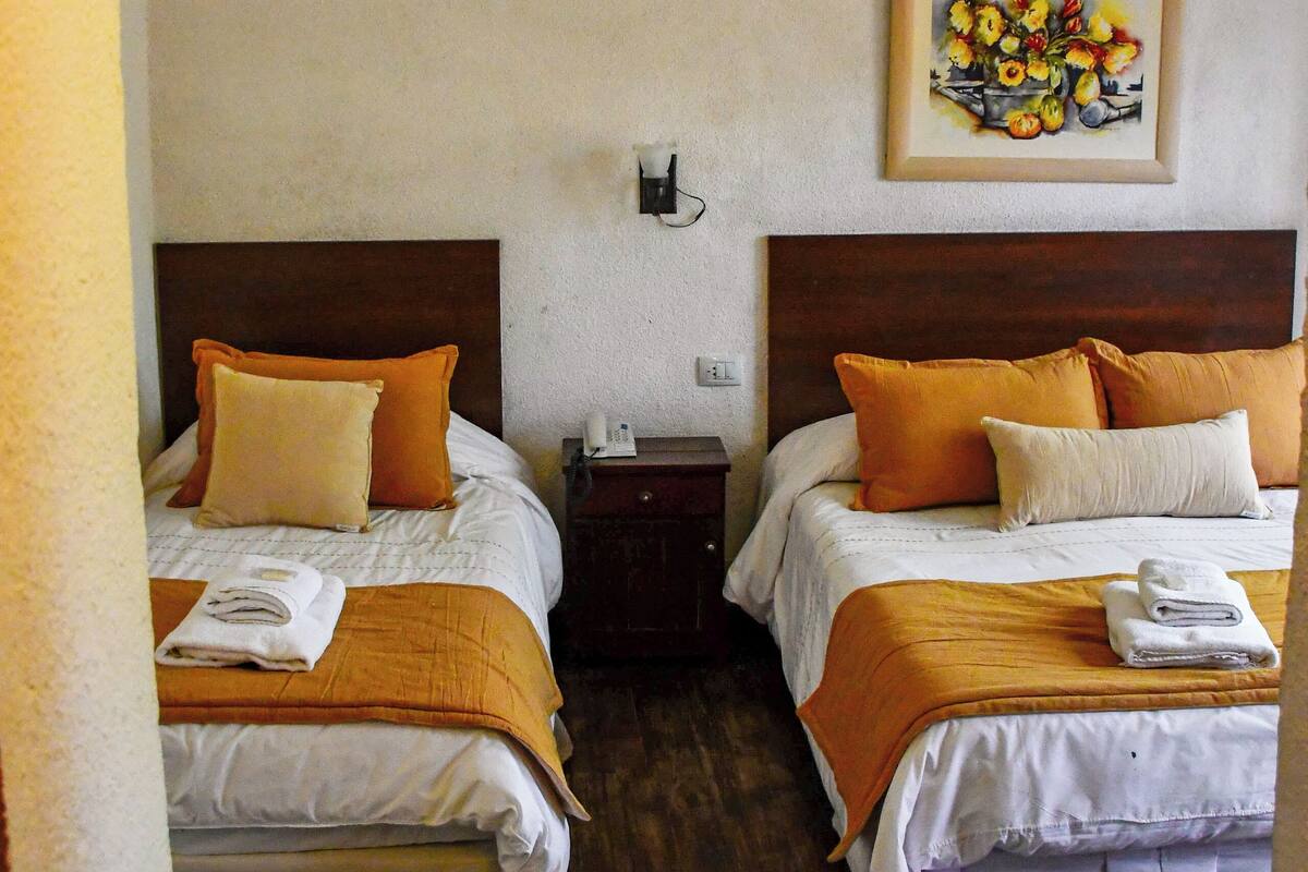 A hotel room with two beds, each with orange and white bedding, a wooden headboard, a nightstand, a painting on the wall, and a wooden floor.