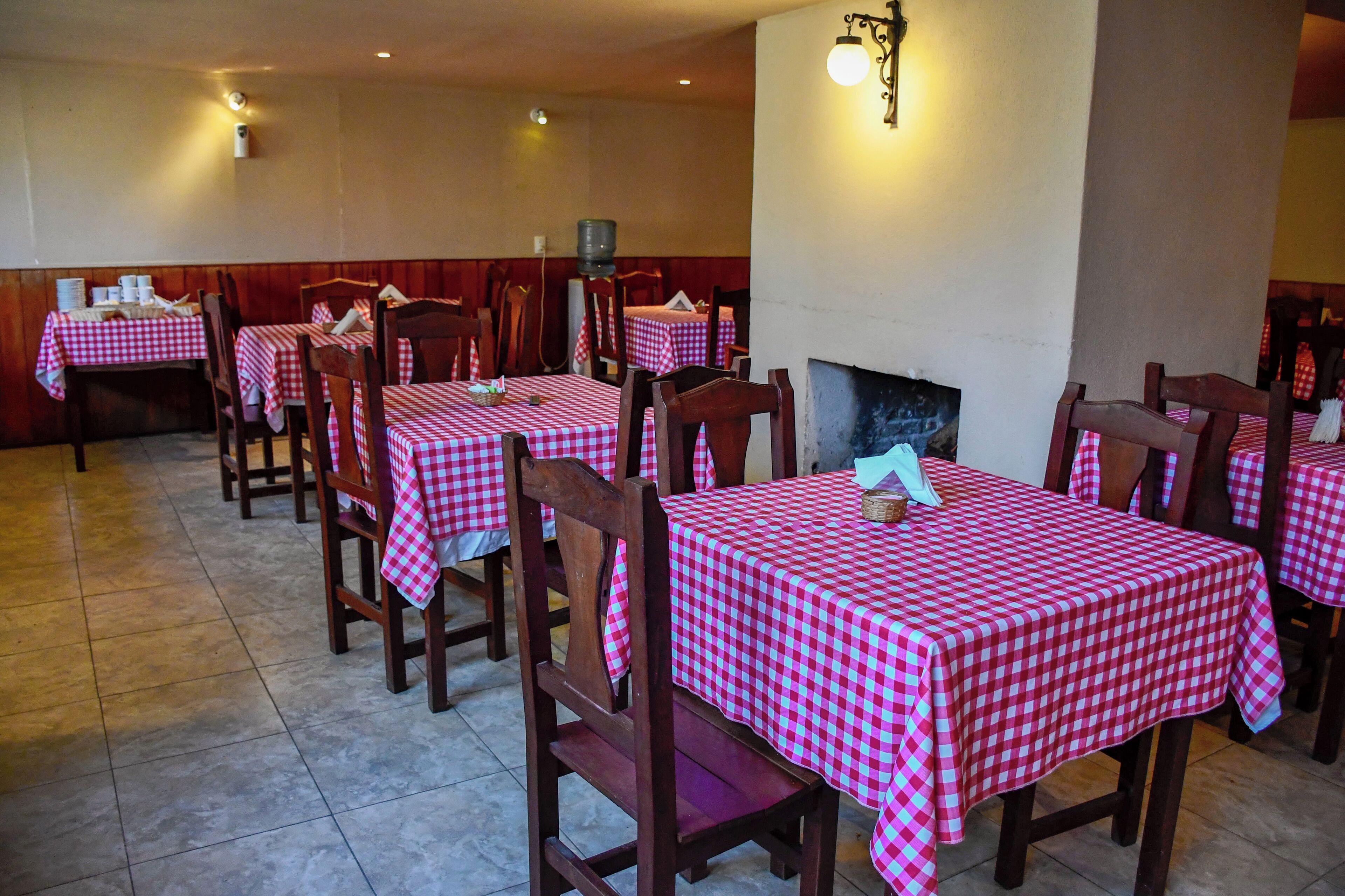 A restaurant with multiple tables covered in red and white checkered tablecloths, wooden chairs, and a tiled floor.
