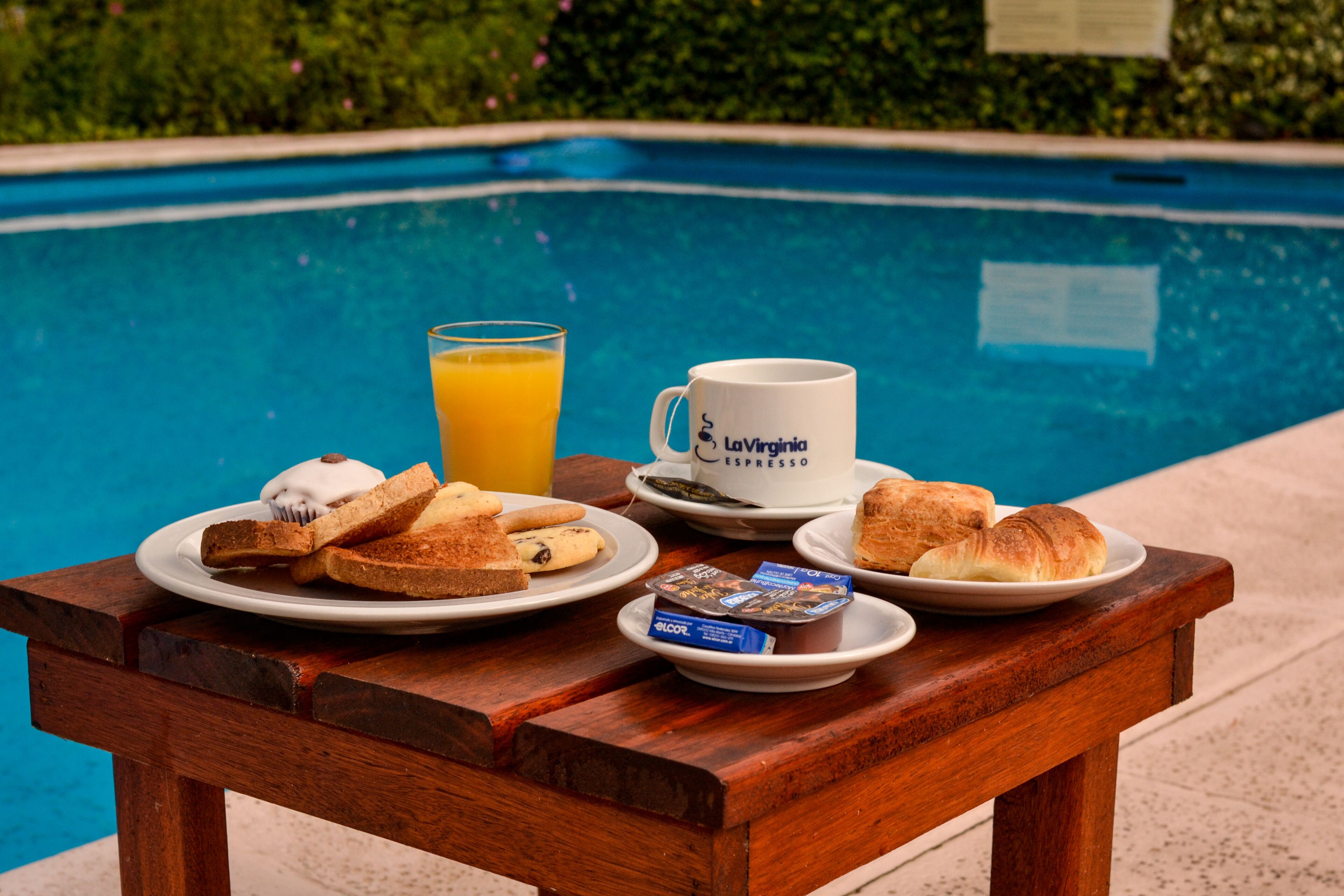 Desayuno junto a la piscina con tostadas, croissants, una taza de café y un vaso de zumo de naranja.