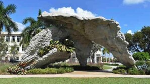 Una gran escultura de roca en un área ajardinada con palmeras y un edificio al fondo.
