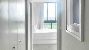 A corridor with a bed at the end, a window with curtains, and a door on the left.
