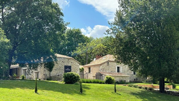 Un bâtiment en pierre, surmonté d’un toit en tuiles, entouré d’une végétation luxuriante et d’arbres.