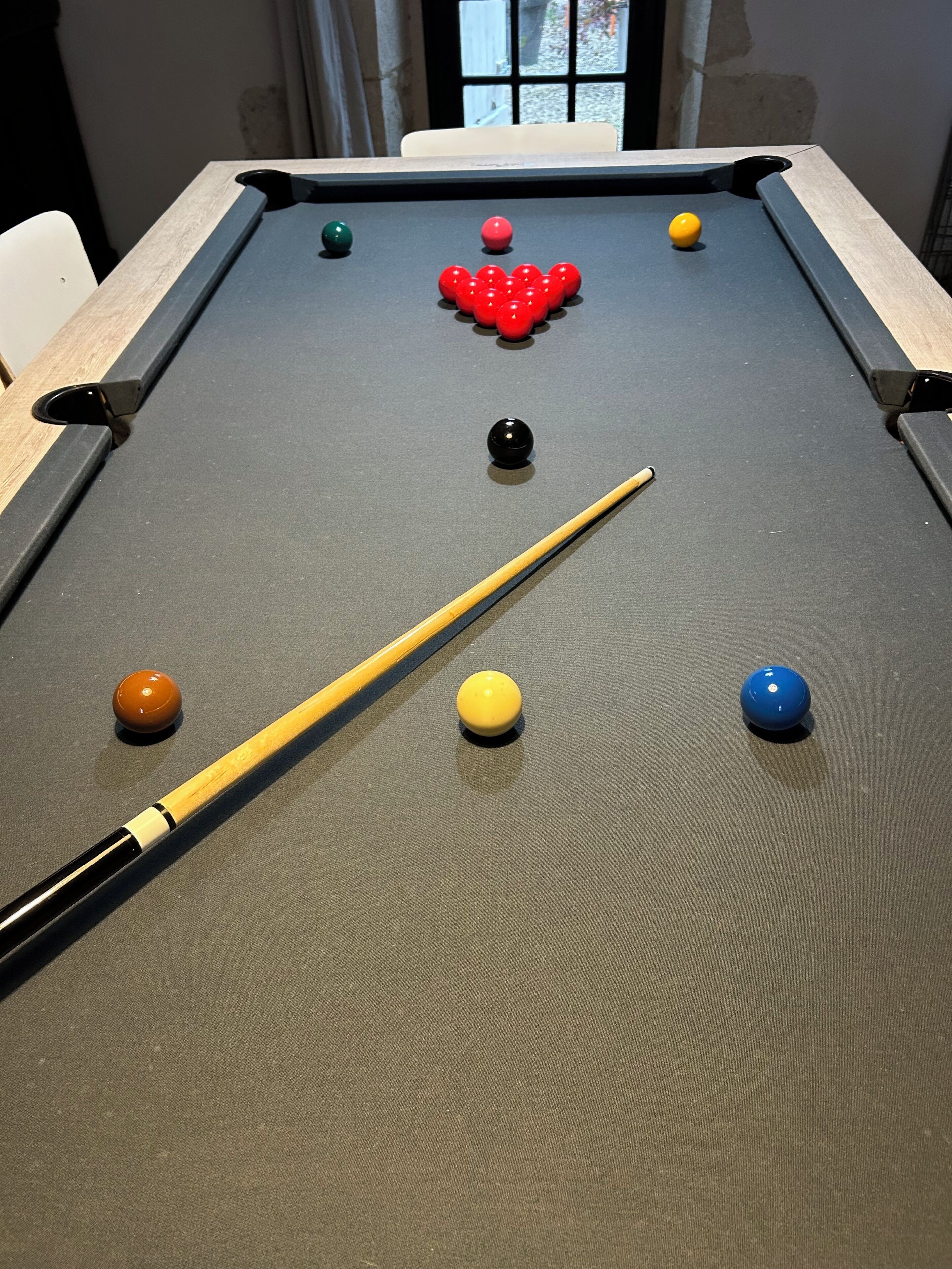 Une table de billard avec les billes disposées en forme de triangle, un queue de billard et une fenêtre en arrière-plan.