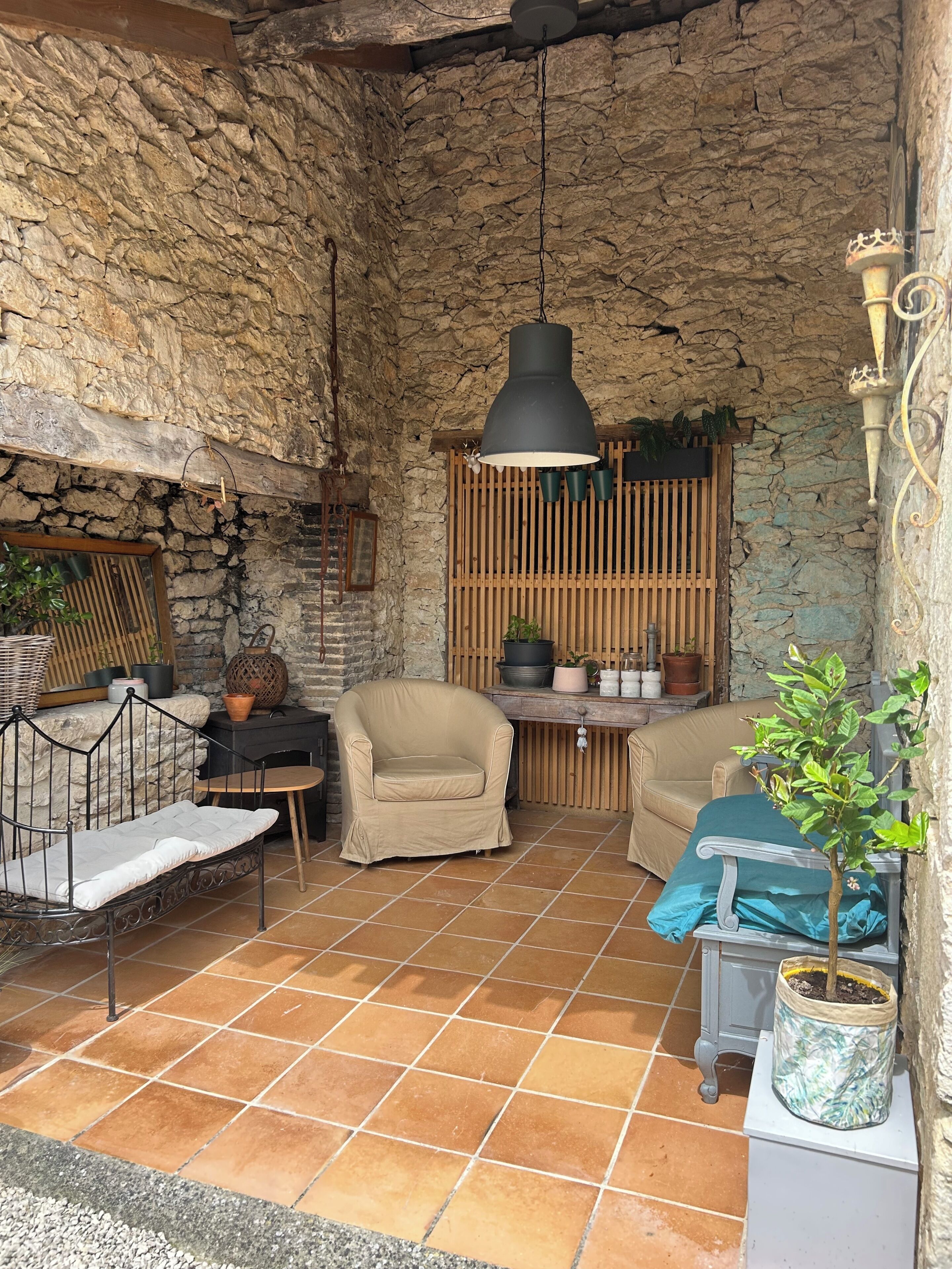 Une terrasse aux murs de pierre, agrémentée d’une lampe suspendue, d’un fauteuil rembourré, d’une petite table et d’une plante en pot.