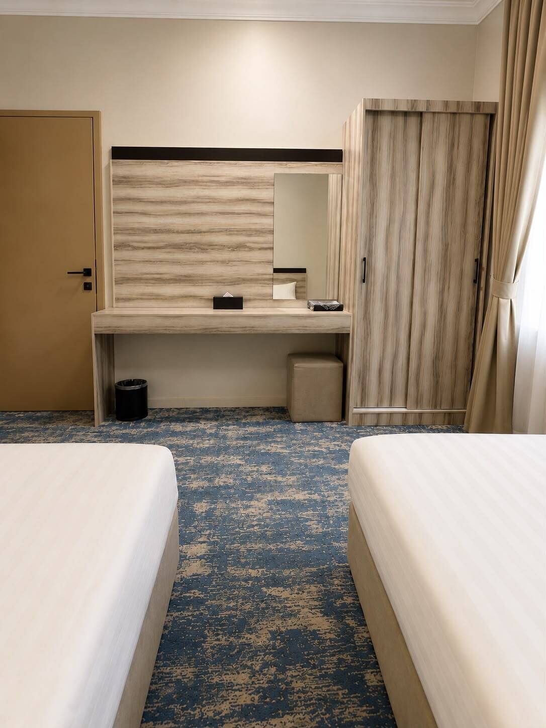 A hotel room with two beds, a desk, a mirror, a wardrobe, and a trash bin.