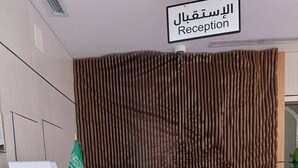 A reception area with a sign indicating 'Reception' in Arabic and English. There is a flag, a framed certificate, and a digital display on the counter. - Tallaq hotel (Madinah)