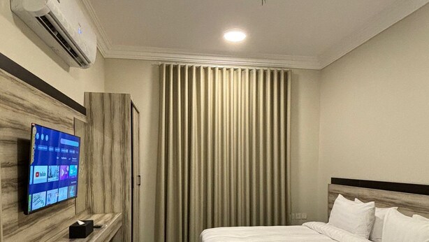 A hotel room with a bed, a television on a wooden shelf, a window with vertical blinds, and a carpeted floor. - Tallaq hotel (Madinah)