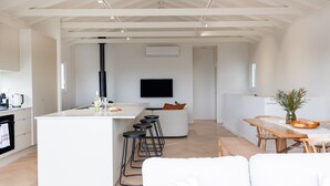 A modern kitchen with a central island, bar stools, a dining table, and a TV mounted on the wall. - Igloo - Whole House in Mollymook (Mollymook)