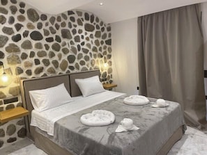 A hotel room with a stone wall, a bed with white linens, two bedside tables with lamps, and a large window with curtains. - Latramys Suites (Naxos)
