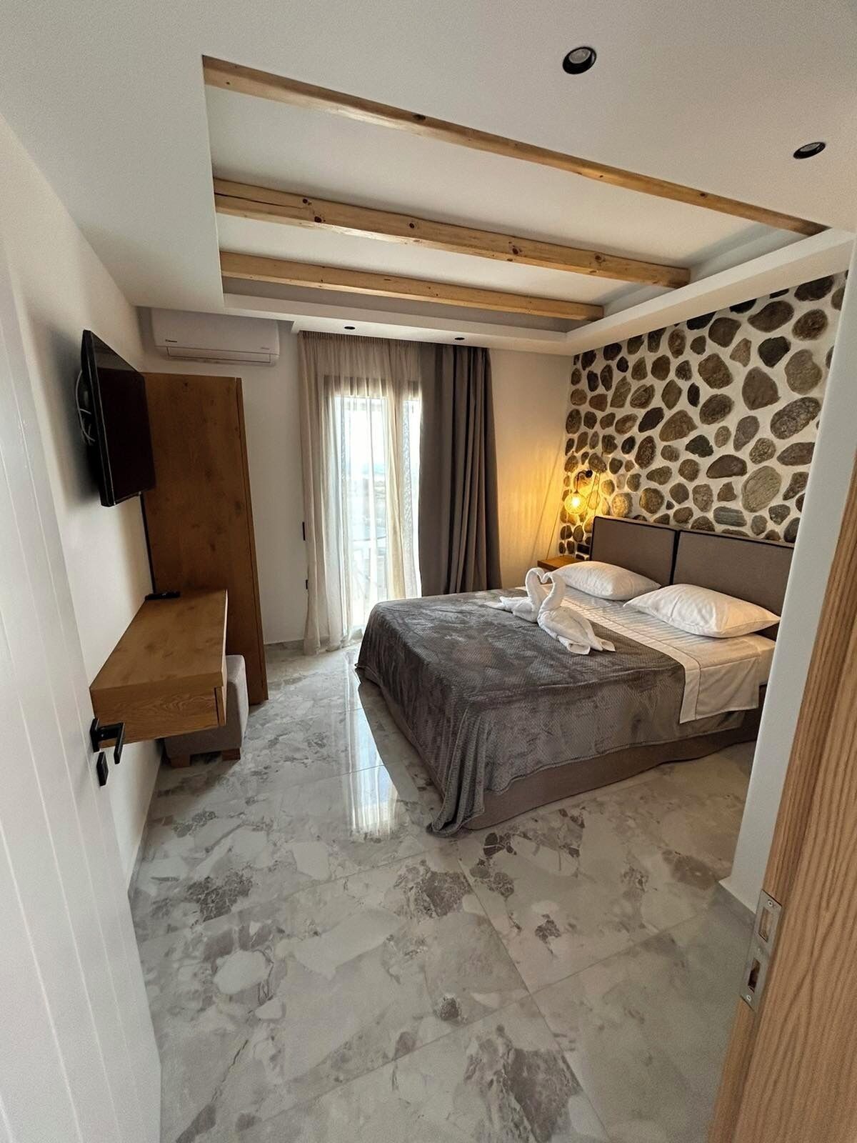 A bedroom with a bed, a stone wall, a wooden shelf, and a TV on the wall.