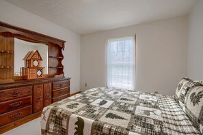 A bedroom with a wooden dresser, a large mirror, a quilted bedspread, and a window with a view of trees.