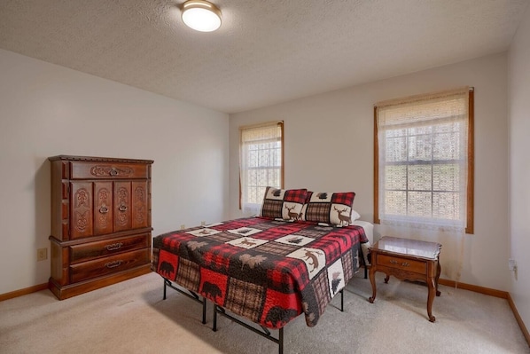 Une chambre à coucher comprenant un lit, une commode, une table de chevet et deux fenêtres ornées de rideaux à motifs.