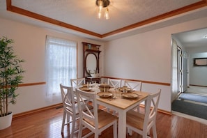 A dining area with a wooden table set for six, white chairs, a potted plant, and a large mirror on the wall.