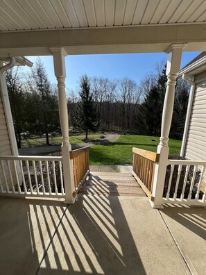 A covered porch with white railings and columns, leading to a grassy yard with trees and a clear sky.