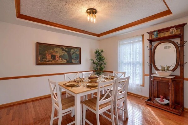 A dining room with a wooden table set for four, white chairs, a painting on the wall, and a wooden cabinet with a mirror and decorative items.