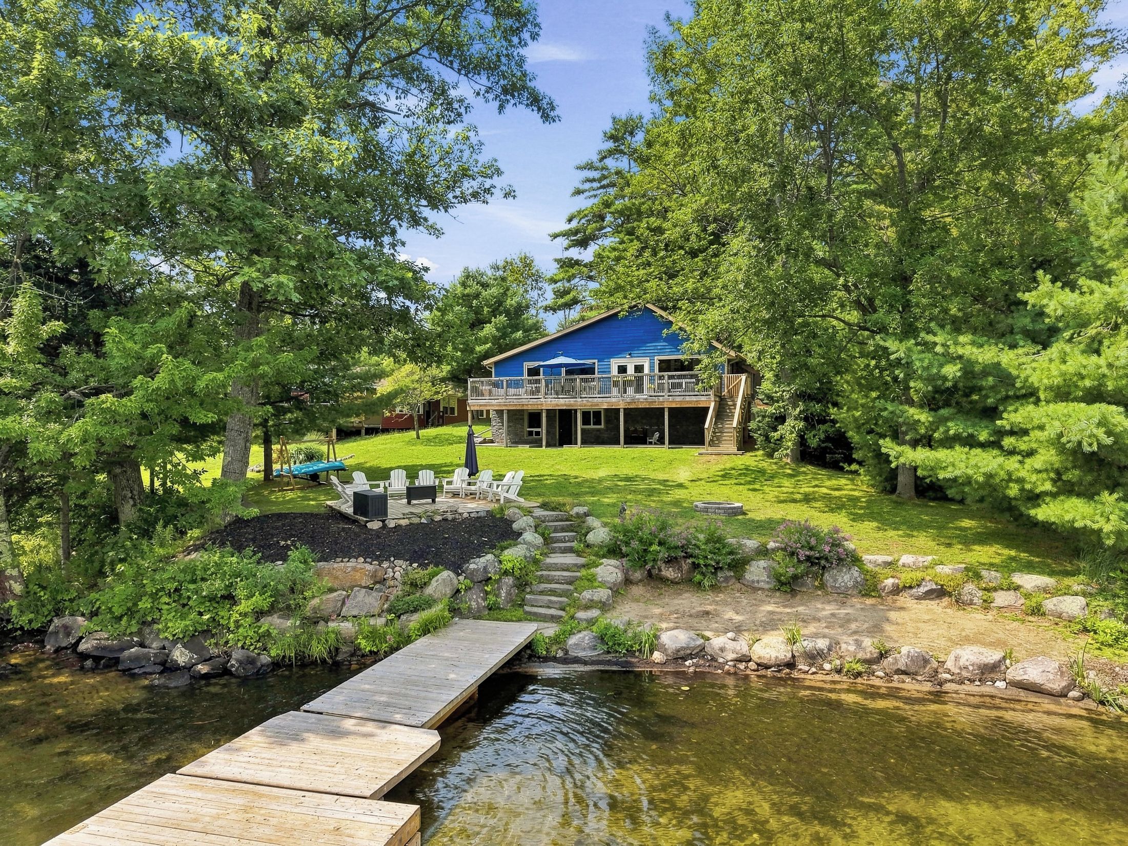 A lakeside cabin with a deck, outdoor seating, and a stone pathway leading to a dock.
