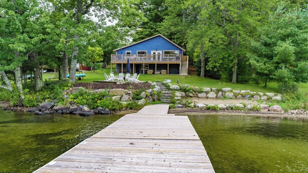 A wooden dock leading to a blue house with a balcony, surrounded by greenery and a pond. - Muskoka Riverside Retreat on the Indian River in the heart of Port Carling (Port Carling)