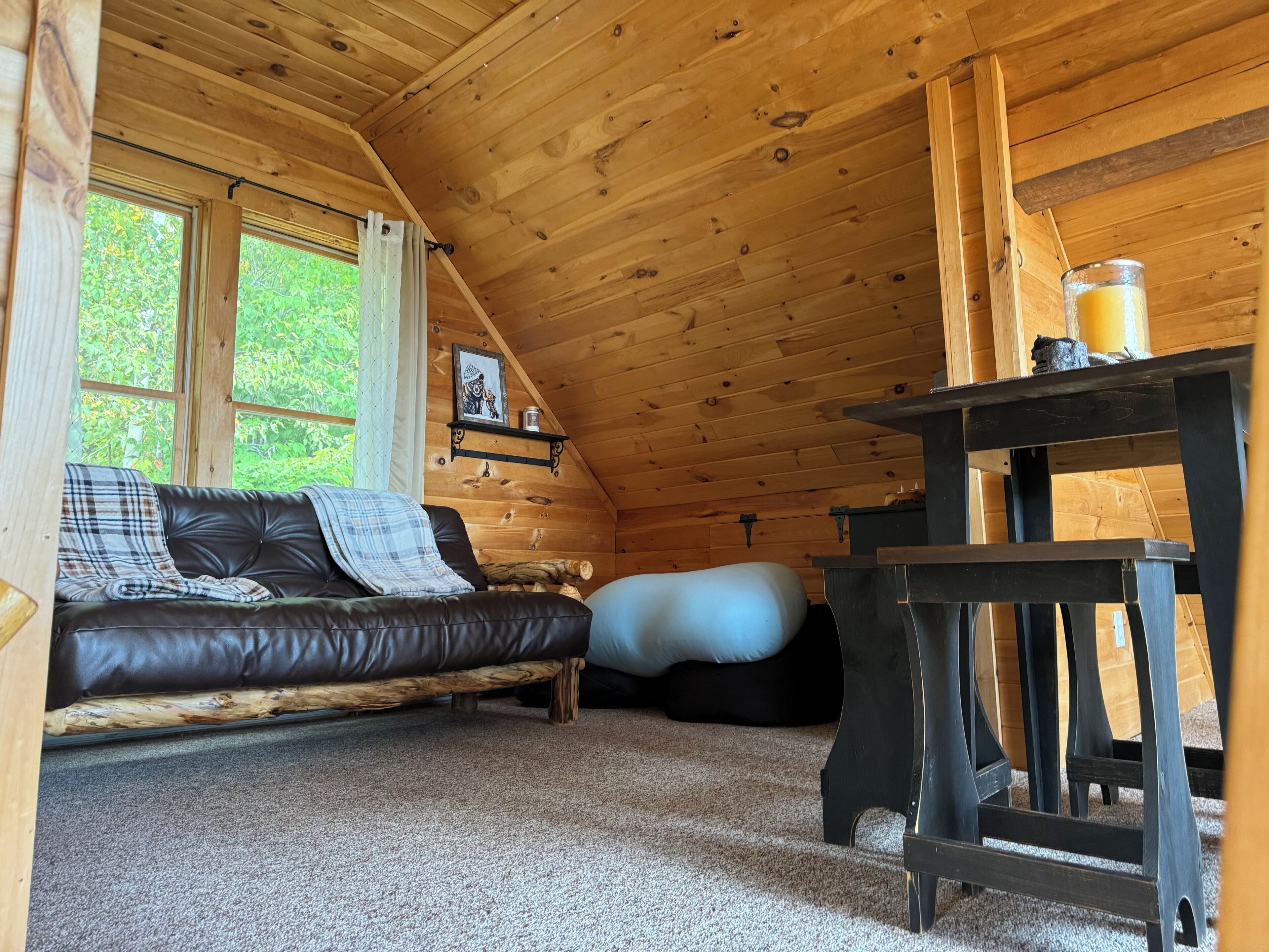 A cozy cabin interior with a leather sofa, a bean bag, a wooden table, and a window with a view of greenery.