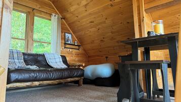 A cozy cabin interior with a leather sofa, a bean bag, a wooden table, and a window with a view of greenery.