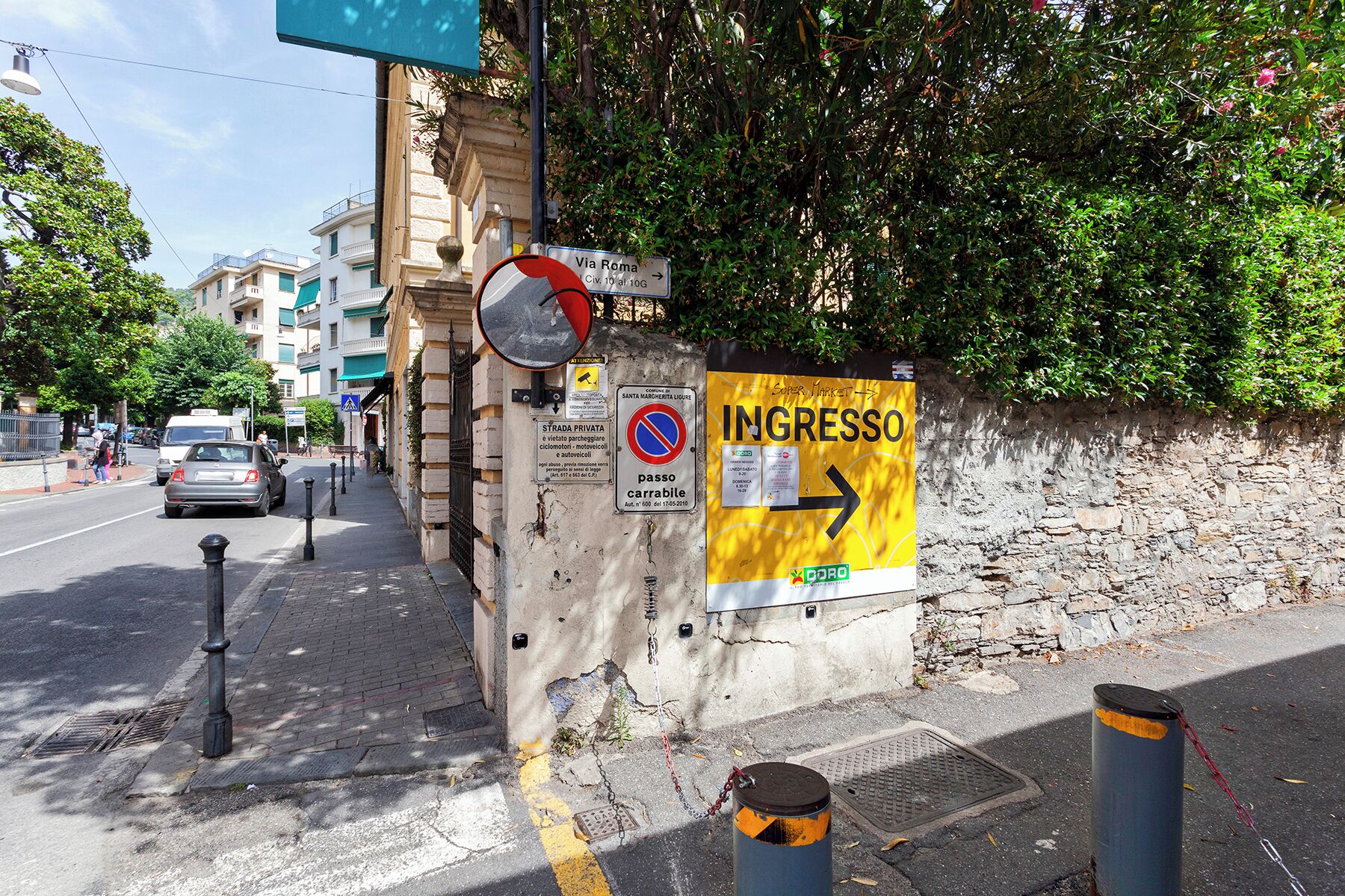 Una esquina de calle con un letrero amarillo que dice 'INGRESSO', una señal de no estacionamiento y un letrero que indica 'Via Roma'.