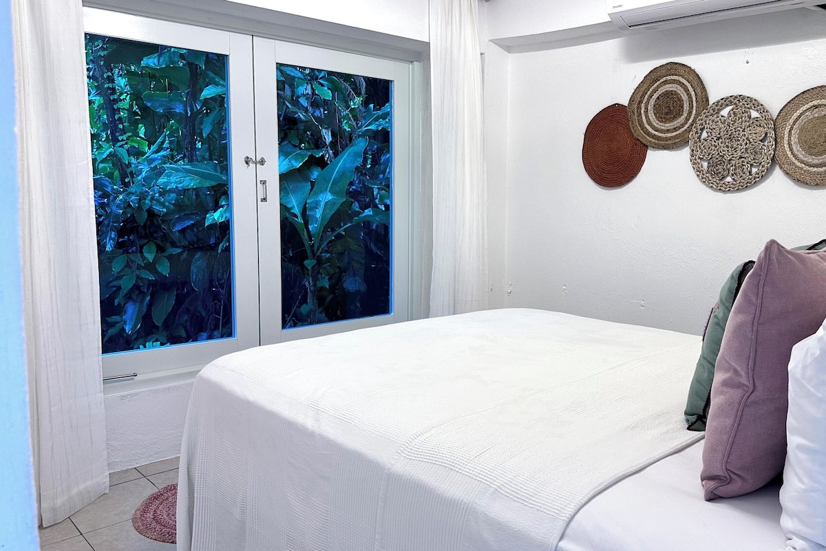 A bedroom with a bed, pillows, a window with a view of foliage, and wall decor.