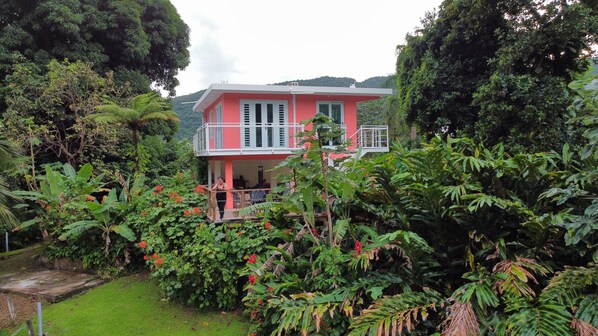 Una casa a due piani con un balcone, immersa nel verde e circondata da piante tropicali.