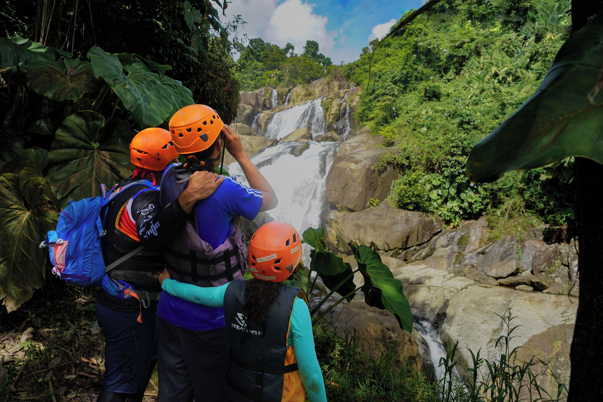 Three people wearing helmets and backpacks, standing near a waterfall surrounded by lush greenery.