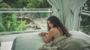A person lying on a bed with a view of a natural landscape through a window. - El Yunque Rainforest (Naguabo)
