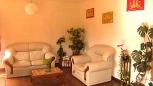 Ένα σαλόνι με έναν μπεζ καναπέ, ένα ξύλινο τραπεζάκι σαλονιού και δύο πολυθρόνες. Στους τοίχους υπάρχουν γλάστρες με φυτά και διακοσμητικά έργα τέχνης.