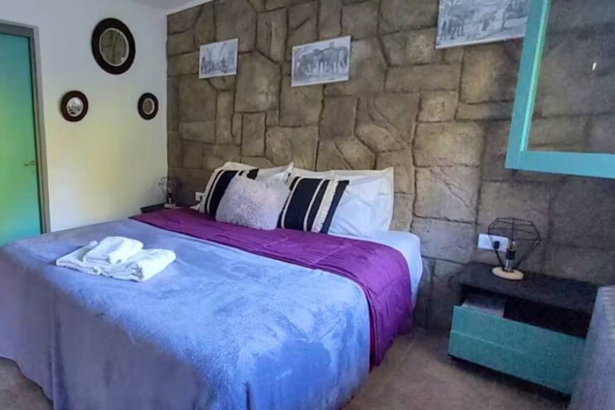 Een slaapkamer met een stenen muur, een bed met een paarse deken en een klein kitchenettegedeelte.