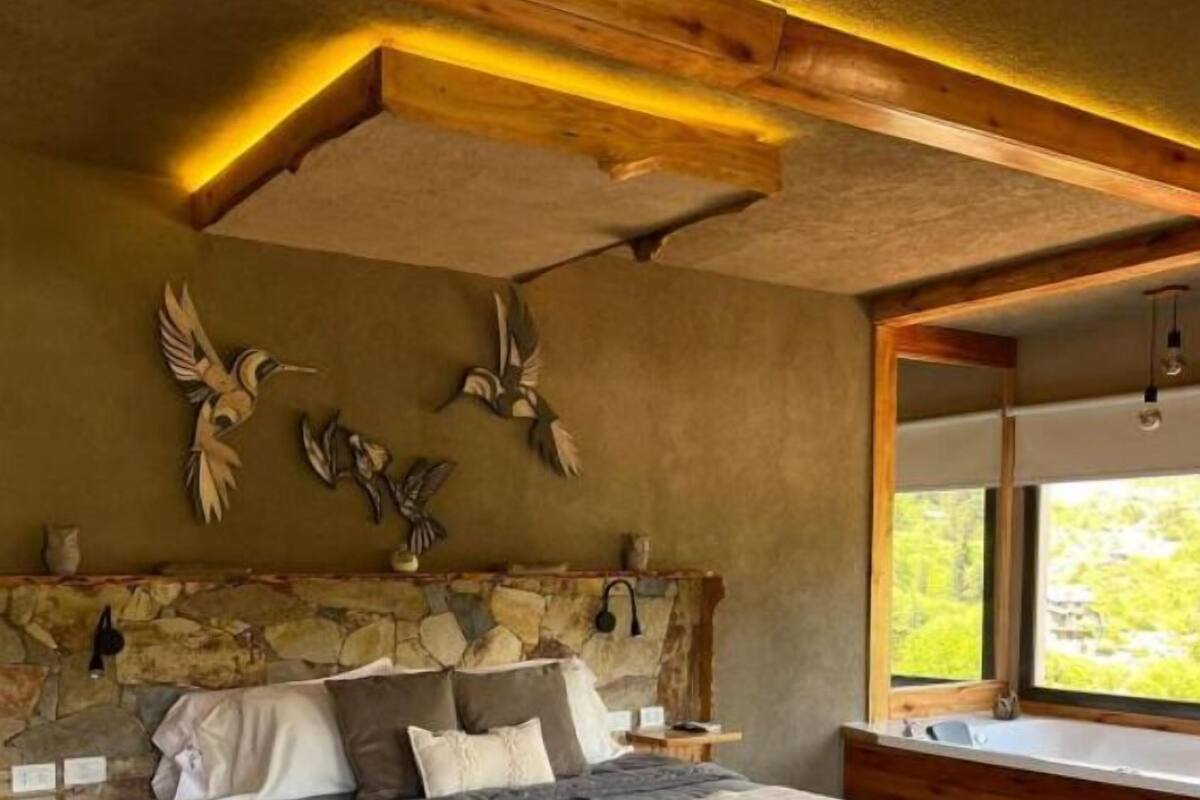 Een slaapkamer met een stenen hoofdbord, een groot bed met veel kussens, een zacht vloerkleed en uitzicht op groen door een raam.
