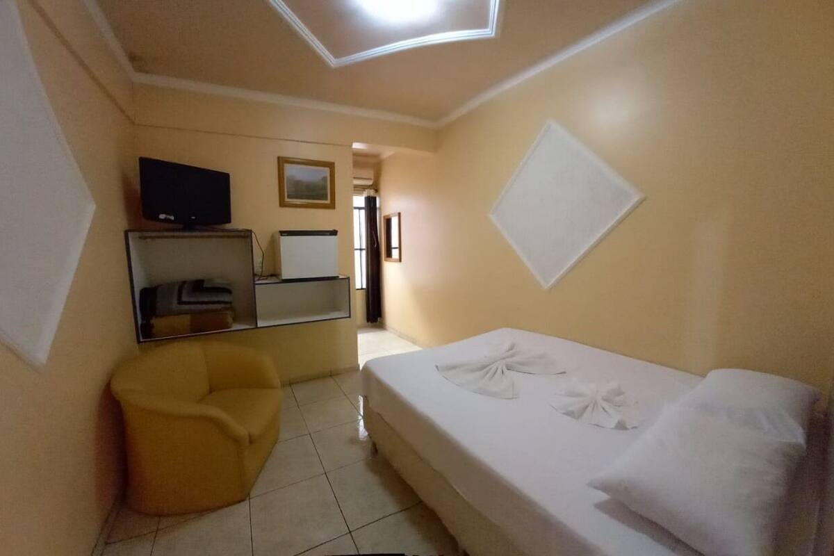 A hotel room with a bed, a TV, a mirror, and a yellow armchair.
