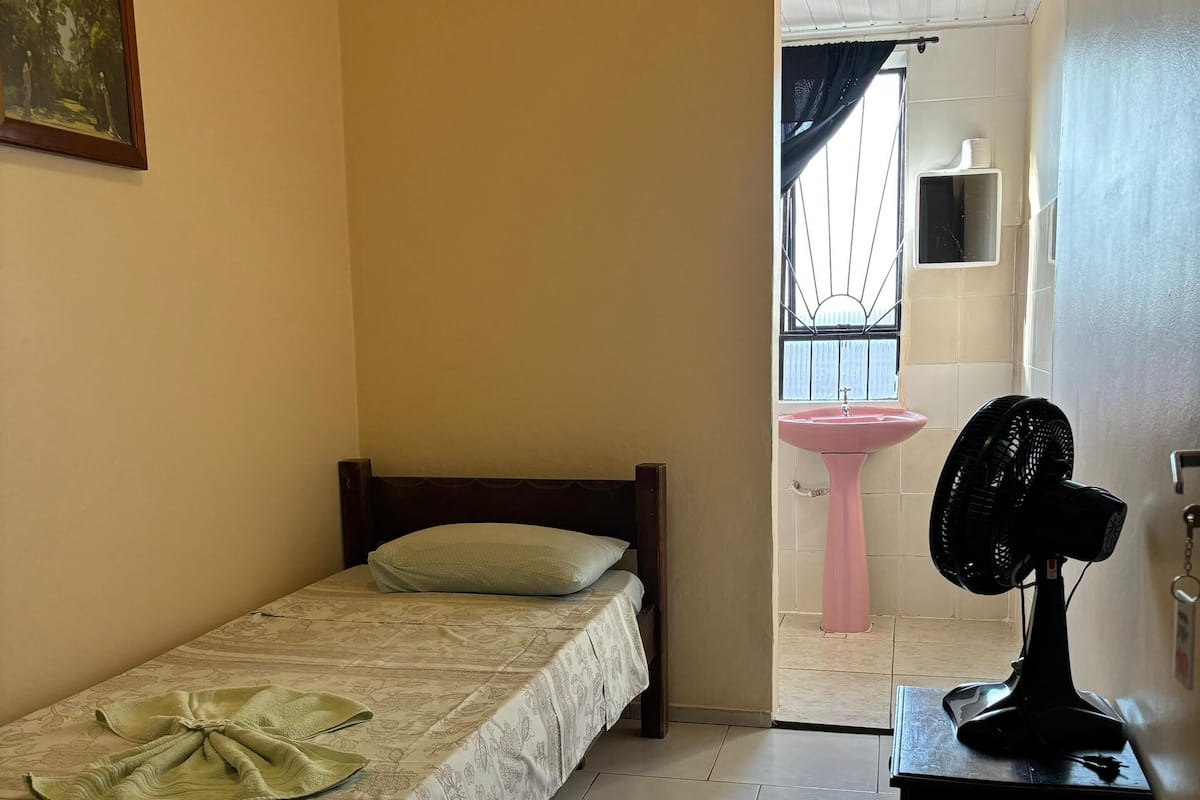 A bedroom with a bed, a bedside table with a fan, a pink sink, and a window with curtains.